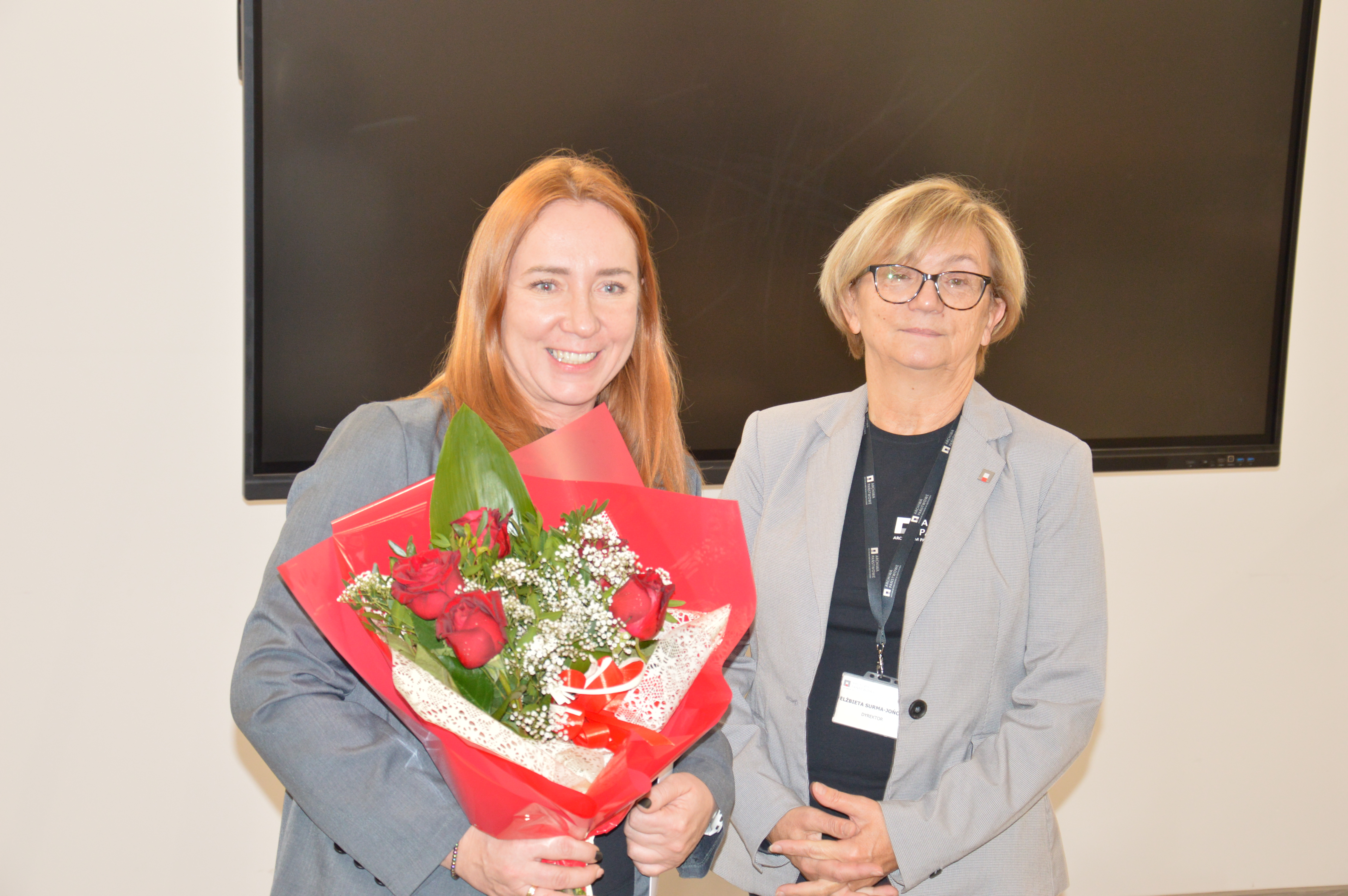 Dwie kobiety stojące przodem. Jedna z nich trzyma bukiet kwiatów.