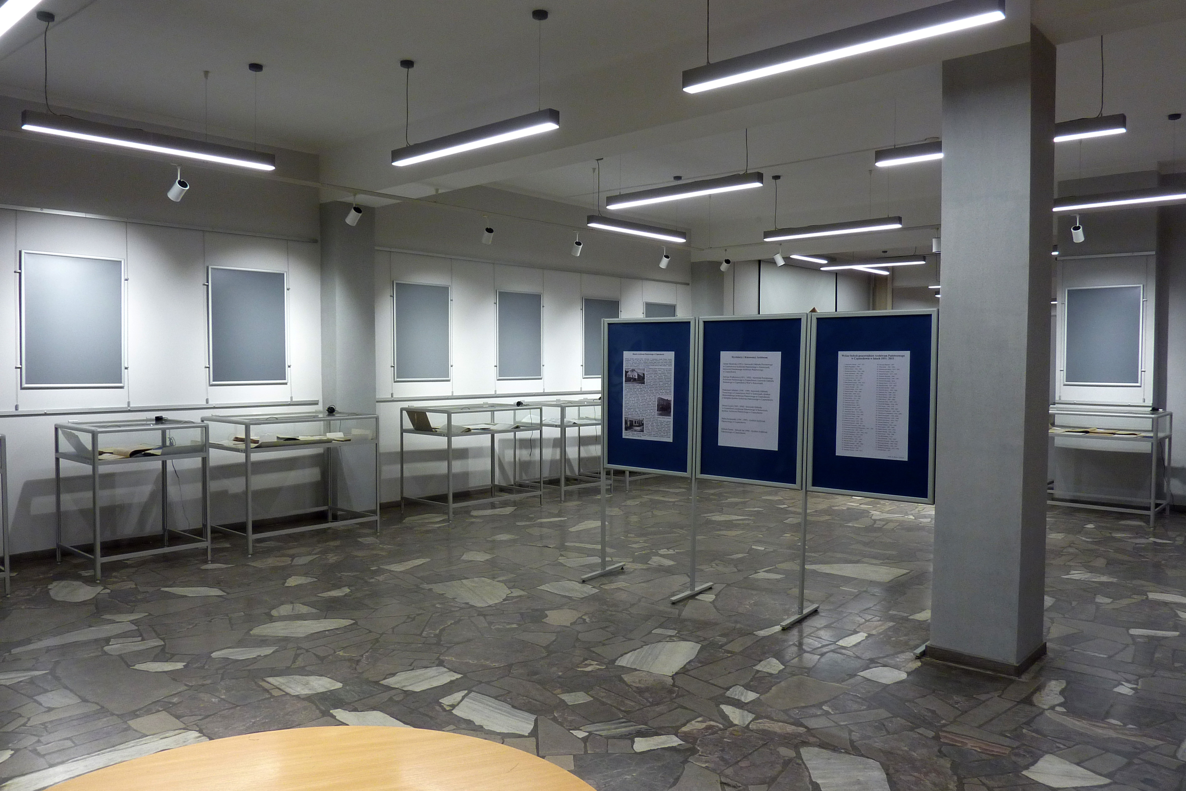 Duża oświetlona sala z gablotami i tablicami do prezentowania materiałów archiwalnych.