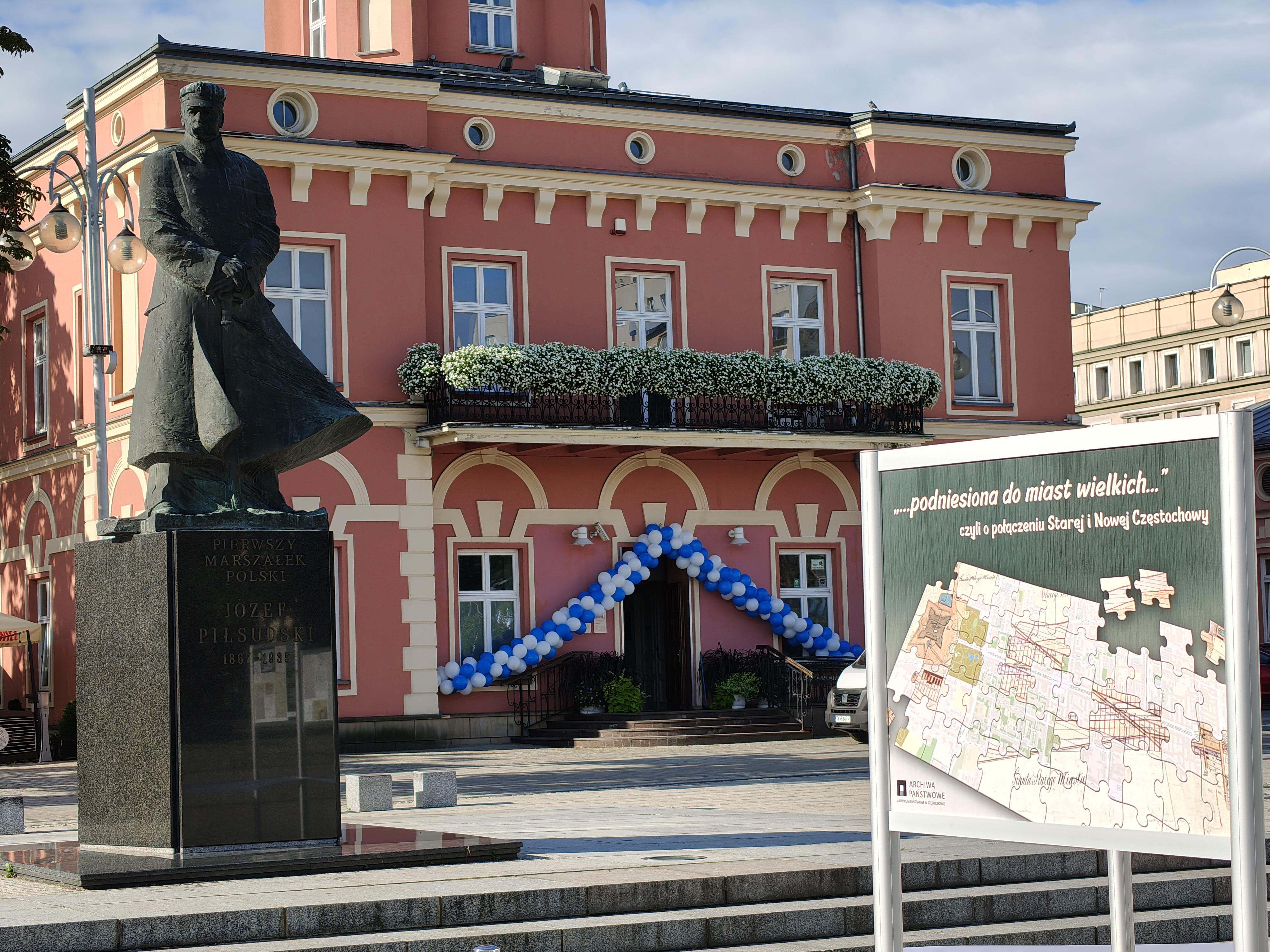 plansza z tytułem wystawy, w tle budynek i pomnik